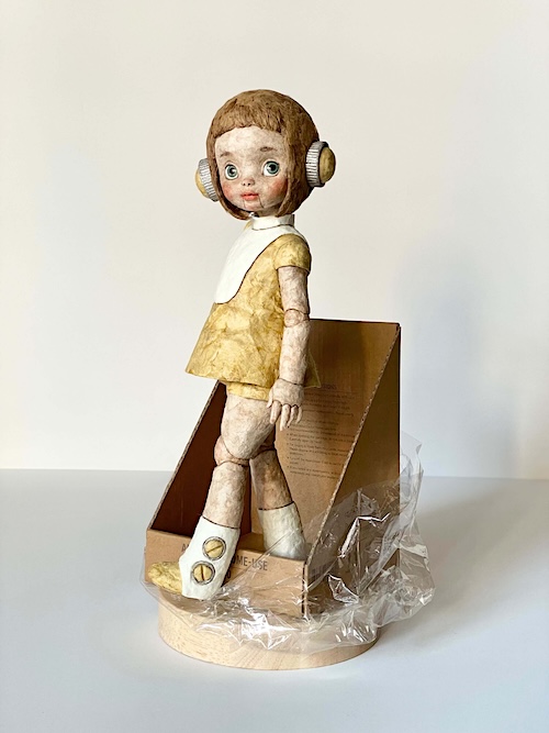 和紙で製作された少女型アンドロイド人形「未来のかたち(ミライちゃん)」の全体像。高さ約31cmの人形が丸い木製台の上に立ち、ダンボールとビニールに囲まれた状態で一歩踏み出すポーズをとっている。